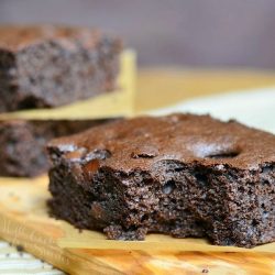 close up view of 2 dark chocolate chocolate chip brownies stacked with wax paper in between each stacked in background with 1 brownie in foreground on a wooden cutting board on a tand and white placemat on wood table