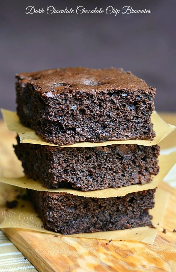 3 dark chocolate chocolate chip brownies stacked with wax paper in between each stacked on a wooden cutting board on a tand and white placemat on wood table
