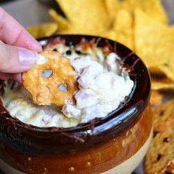 close up view of hand pulling out hot asiago ham dip portion on a pretzel chip out of a brown and tan crock with pretzels and chips scattered around base of crock while all sit on a blue cloth