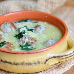 close up view of Orange and tan clay crock with Italian Wedding Soup with turkey meatballs and Orzo sitting on a tan table cloth on a wood table