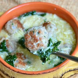 above view of Orange and tan clay crock with Italian Wedding Soup with turkey meatballs and Orzo sitting on a tan table cloth on a wood table with a spoon picking up a meatball