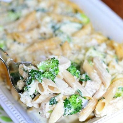 White rectangular baking dish with Lightened spinach broccoli chicken alfredo bake on a white and green decorative placemat on a wooden table and a spoon holding one portion of the bake