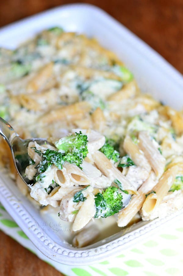 White rectangular baking dish with Lightened spinach broccoli chicken alfredo bake on a white and green decorative placemat on a wooden table and a spoon holding one portion of the bake
