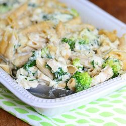 Close up view of White rectangular baking dish with Lightened spinach broccoli chicken alfredo bake on a white and green decorative placemat on a wooden table and a spoon holding one portion of the bake