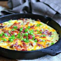 close up view of Black skillet with loaded baked potato breakfast on grey cloth sitting on wood table garnished with green onion