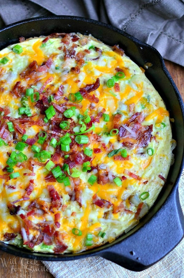 Loaded Baked Potato Breakfast Skillet appears in a black skillet. There is cheese, chopped bacon and green onions on top of the cooked egg and potato mixture.