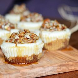 close up view of 6 pecan mini cheesecakes lined up in 2 rows on a wood cutting board with maple syrup drizzled across the tops of the cupcakes