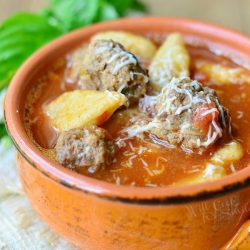 close up of orange crock filled with spicy meatball gnocchi soup on a white placemat on wood table with leaf in background