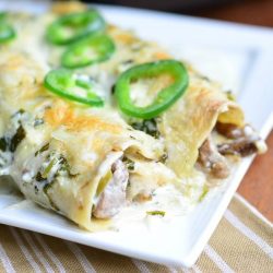 close up view of 2 steak enchiladas with jalapeno cilantro cream sauce on a white rectangle plate on a tan and white cloth on a wood table