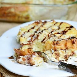 close up view of one portion of almond joy french toast bake on a round white plate on tan placemat with baking dish and rest of bake in background and fork taking piece of french toast