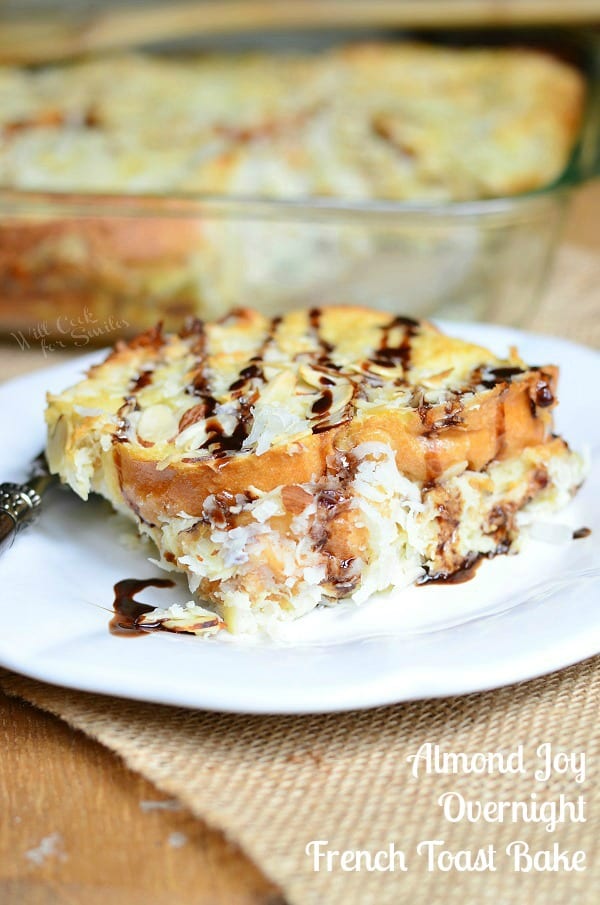 One piece of Almond Joy French Toast Bake served on a white plate. Chocolate sauce is drizzled over the top. The casserole dish fades into the background.