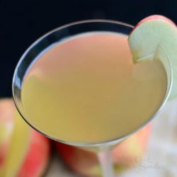 close up view from the top down of a martini glass with apple cider martini on a tan tablecoth and apples below the glass itself.