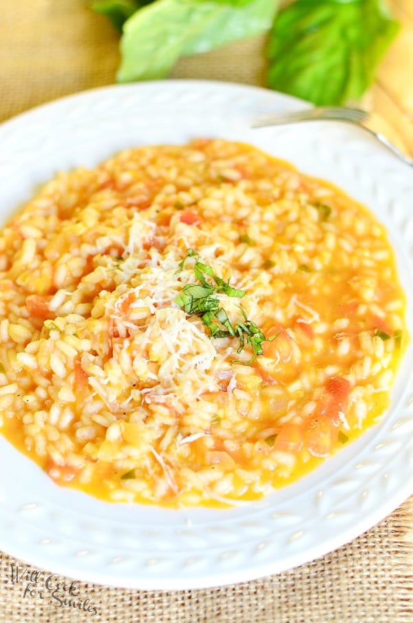 view from above of a white decorative rimmed bowl filled with creamy tomato basil risotto on a tan placemat on wooden table with leaves of basil as a garnish