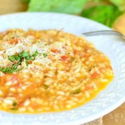 close up veiw of a white decorative rimmed bowl filled with creamy tomato basil risotto on a tan placemat on wooden table with leaves of basil as a garnish