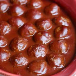 Red ceramic baking dish filled with cooked meatballs in sauce on a tan placemat