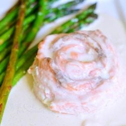 close up and viewed from above of white square plate with salmon filet molded into a rose design in white sauce with asparagus on a tan cloth on wood table with a sauce cup in background