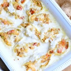 white baking dish filled with seafood alfredo stuffed shells on a tan placemat on wooden table with a silver serving spoon at bottom left of baking dish
