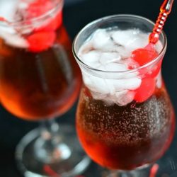 clsoe up view of 2 decorative wine glasses filled with "The pink lady raspberry cake coctail" on a black table with cherries at base of glass