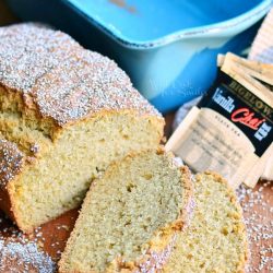 2 slices of vanilla chai tea bread cut from loaf laying on a wooden table with the rest of loaf behind and a blue baking dish in background with tea bags laying on tan cloth