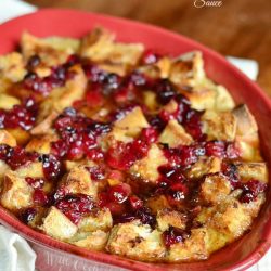 red clay baking dish filled cranberry bread pudding with honey whiskey maple sauce on a white cloth on wood cutting board