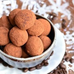 close up view of decorative white bowl filled above the brim with espresso chocolate truffles on a white plate with coffee beans strewn about the bottom while all sit on a wooden table