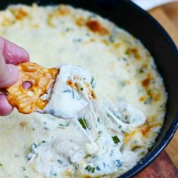 hand holding pretzle chip with one bite of dip being held above a black skillet with four cheese hot dip on a wooden table with a brown table cloth in the background