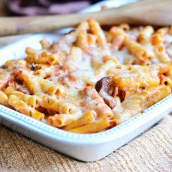 close up view of white baking dish with Itlian cheese steak baked ziti on a tan placemat with a wooden spoon spanning the corner of dish