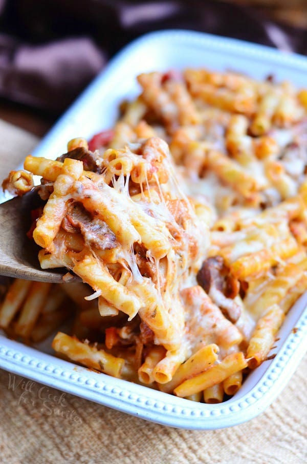 Italian Cheese Steak Baked Ziti is served in a white casserole dish. A wooden spoon lifts some of the pasta up from the dish. Pasta, meat and cheese are the main components here.