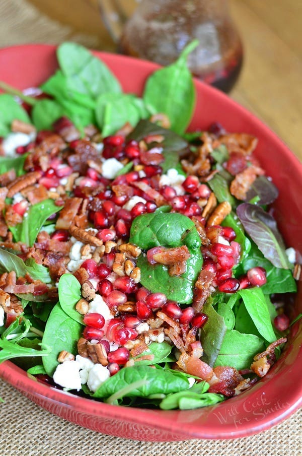 pomegranate goat cheese salad in a red baking dish on a tan placemat on wooden table with a bottle of dressing in background to the left
