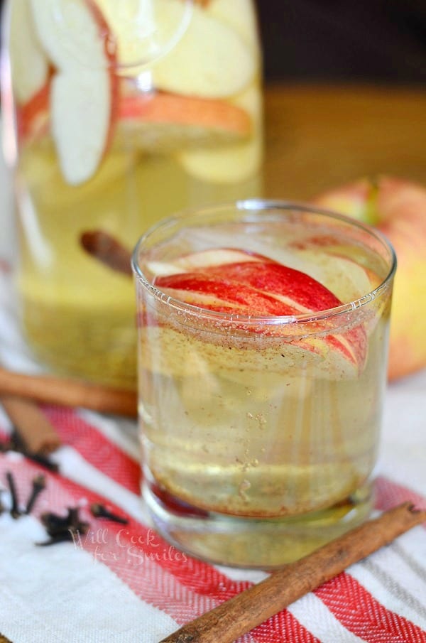 small glass filled with spiced apple sangria with sliced apples floting near the top of the glass while the glass is on a white and red cloth and a cinnamon stick rests at bottom of glass. A glass pitcher rests in background filled with spiced apple sangria and an apple to it's right.