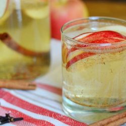 close up view of small glass filled with spiced apple sangria with sliced apples floting near the top of the glass while the glass is on a white and red cloth and a cinnamon stick rests at bottom of glass. A glass pitcher rests in background filled with spiced apple sangria and an apple to it's right.