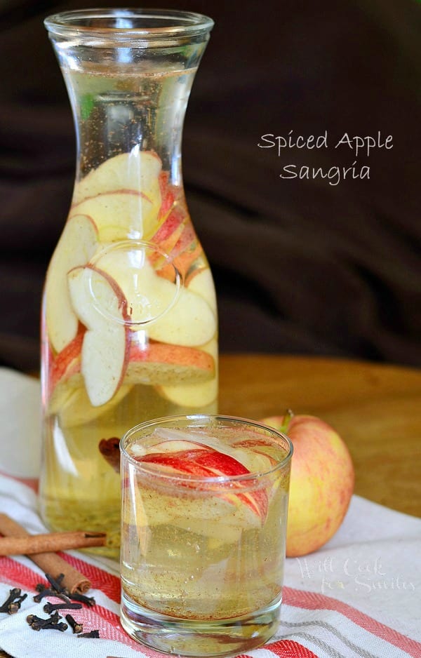 There is a glass and a pitcher of Spiced Apple Sangria with floating red apples. Sticks of cinnamon and another whole apple are also sitting on the tablecloth.