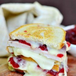 stacked halves of a turkey cranberry brie grilled cheese on a wooden table with a tan cloth in background and a bowl of cranberry sauce in the background to the right