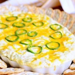 decorative white bowl filled with cheesy jalapeno dip centered in between crackers laying on a silver platter with a white and gold cloth in the background