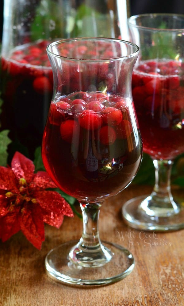 2 wine glasses filled with cranberry sangria on a wooden table in front of a pitcher filled with additional sangria and winter floral decor on table with around the glasses.
