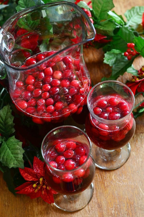 view from above of 2 wine glasses filled with cranberry sangria on a wooden table in front of a pitcher filled with additional sangria and winter floral decor on table with around the glasses.