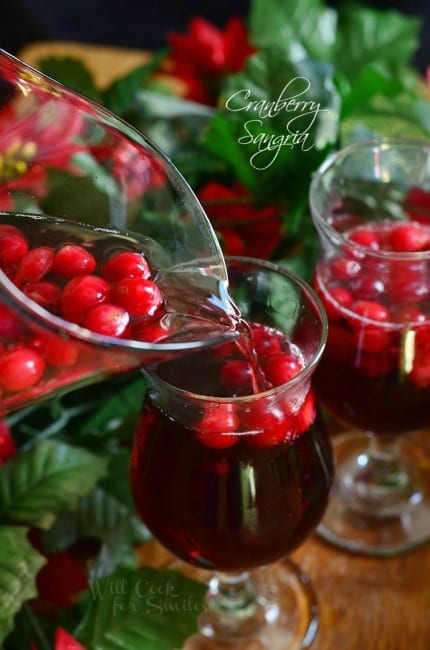 2 wine glasses filled with cranberry sangria on a wooden table as a pitcher filled with additional sangria is being poured into first glassand winter floral decor on table with around the glasses.