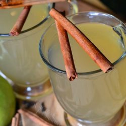 view from above of 2 glass mugs filled with crock pot mulled green apple cider and 2 cinnamon sticks laid across each glasses top rim as they both sit on a wooden table with more sticks at bottom of glasses.