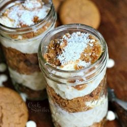 3 mason jars filled with no bake white chocolate cheesecake gingerbread trifles on a wooden table with white candy scattered on table