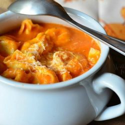 white ceramic crock filled with roasted bell pepper tortellini soup on a wood cutting board with a fall festive cloth in the background with a spoon resting on the crock