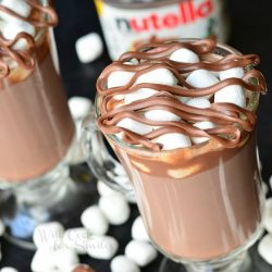 view from above of 2 dessert coffee mugs filled with spiked nutella hot chocolate with mini marshmellows scattered around the base of the glasses and a spoon with a scoop of nutella in foreground as a jar of nutella rests in the background.