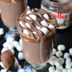 2 dessert coffee mugs filled with spiked nutella hot chocolate with mini marshmellows scattered around the base of the glasses and a spoon with a scoop of nutella in foreground as a jar of nutella rests in the background as viewed from above