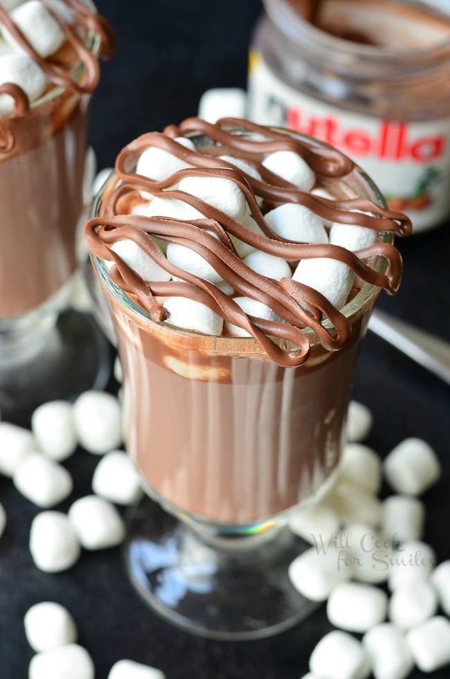 Hot Chocolate in a glass with marshmallows and Nutella on top on a table with mini marshmallows around it