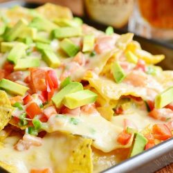 Close up view of Nachos with veggies and homemade nacho ale cheese on a silver baking pan on a wooden table with beer in the background