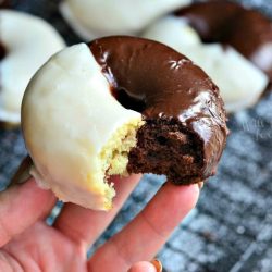 hand holding 1 donut with a bite taken out above Black and white glazed donuts on a cooling rack on a black table with powdered sugar on table