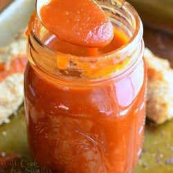 mason jar filled with homemade whiskey BBQ sauce standing on a baking pan with easy baked chicken strips and a spoon lifting some sauce above the jar