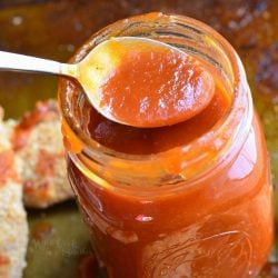 mason jar filled with homemade whiskey BBQ sauce standing on a baking pan with easy baked chicken strips and a spoon lifting some sauce above the jar