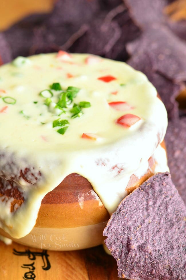 Homemade White Queso Dip that is over flowing in a brown bowl with green onions on top as a garnish and purple tortilla chips around it 