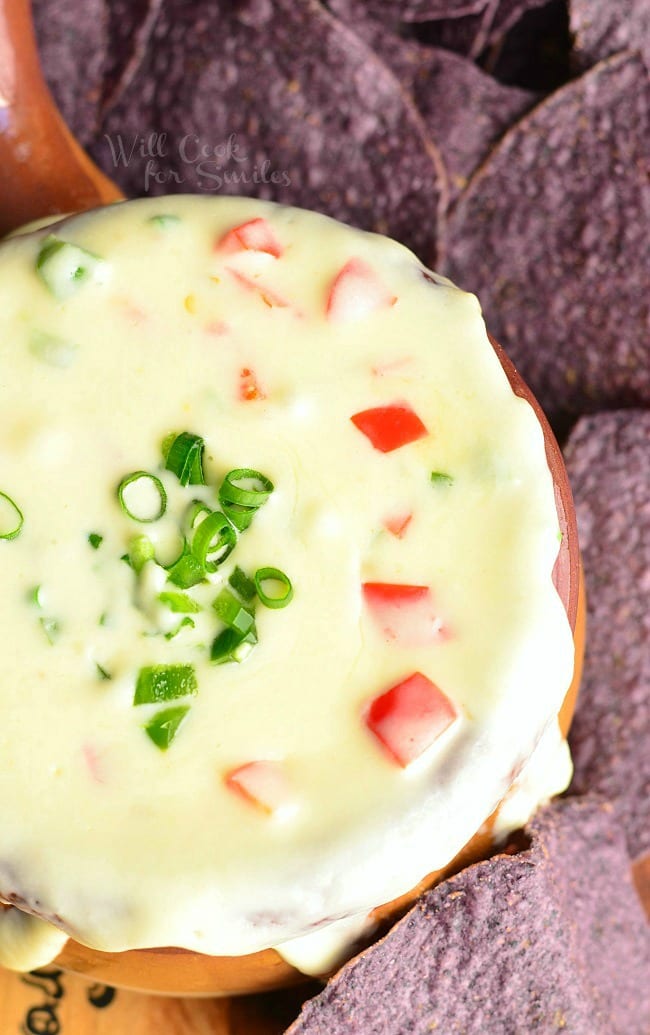 view from above of homemade white queso dip in a brown crock with blue tortilla chips around crock. Queso is topped with green onion