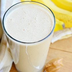 view from above of Pint glass with skinny almond banana breakfast smoothie on a wood table with a spoon and peanut butter at the bottom left, bananas in the background to the right and a white cloth in the background to the left